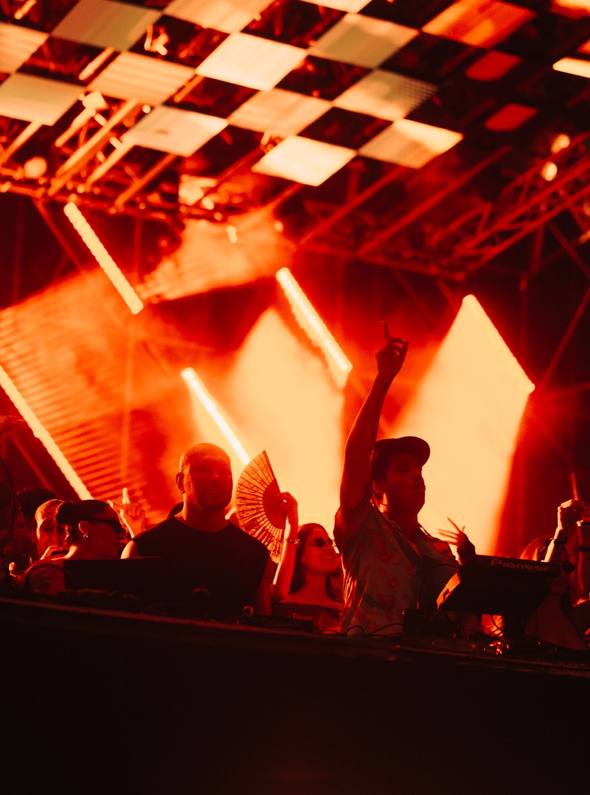 Close view of the DJ booth under the checkerboard stage roof, red stage light on the crowd and performers