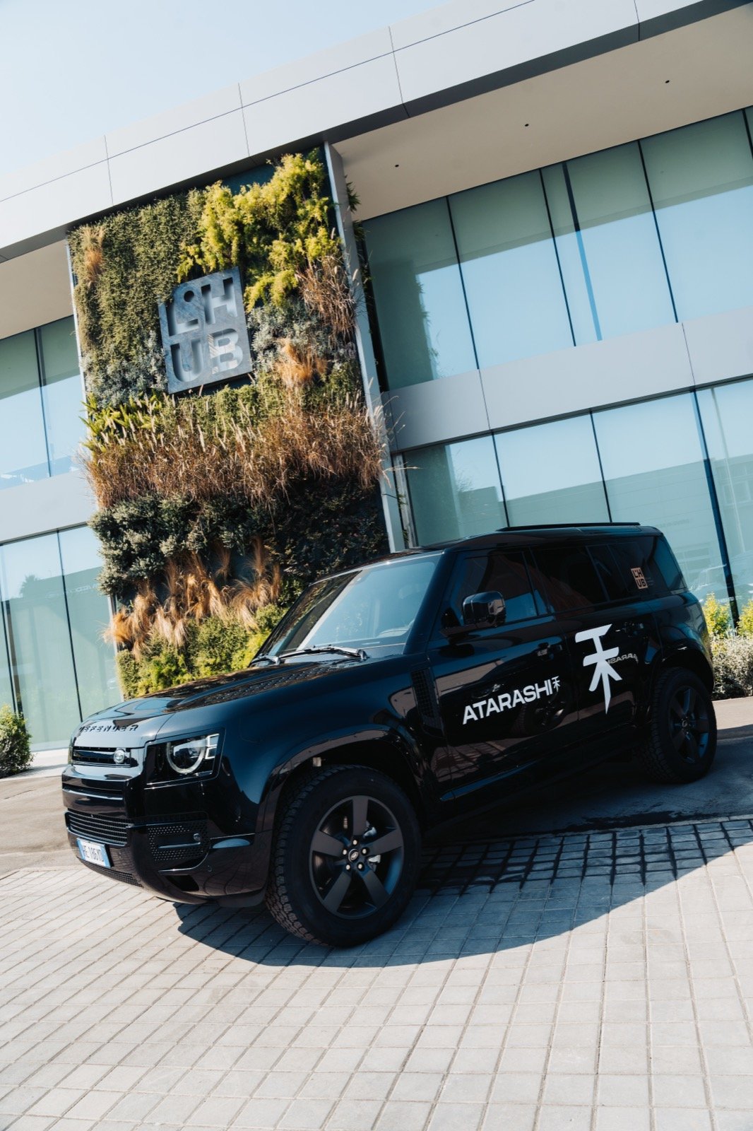 Black Atarashi-branded Land Rover Defender parked outside the L'HUB building, vertical garden and L'HUB signage behind it