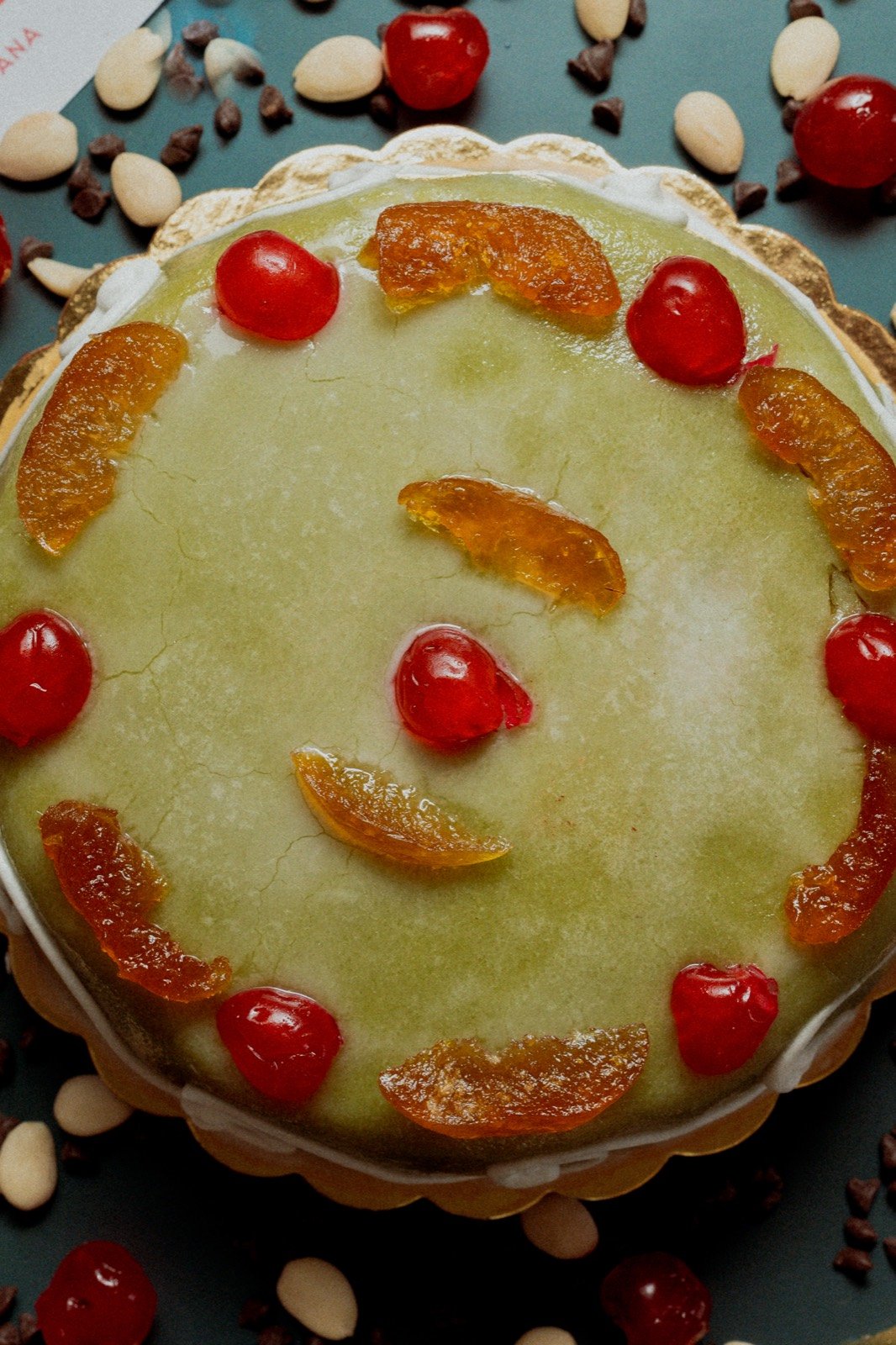 Overhead of a Casa Irrera pastry with pale green icing, candied citrus and glace cherries on a teal surface