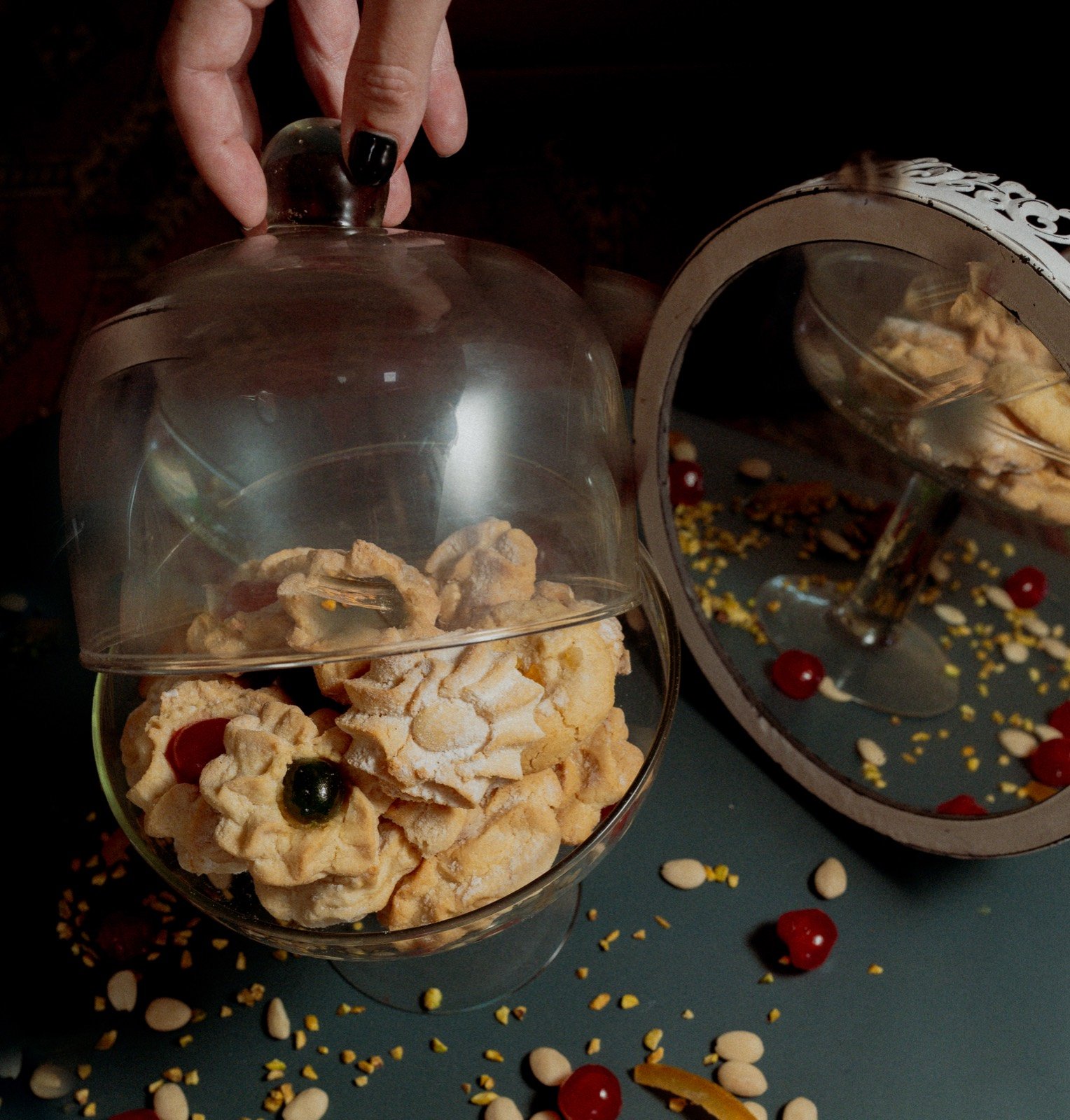 A hand lifting a glass cloche off a bowl of amaretti cookies, mirror reflection beside it