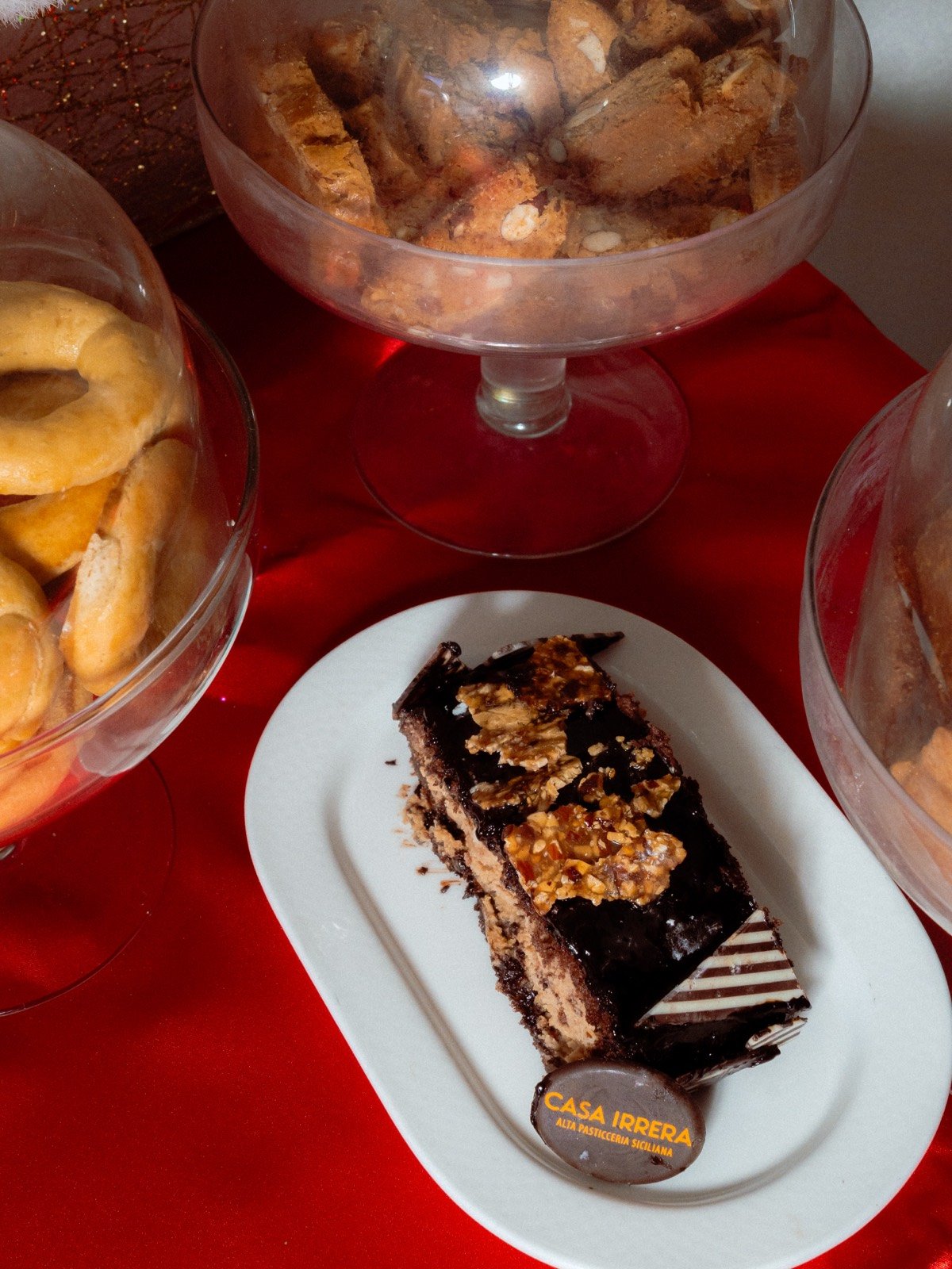 Overhead glass bowls of cantucci biscuits beside a slice of chocolate hazelnut cake with a Casa Irrera branded disc, on a red surface