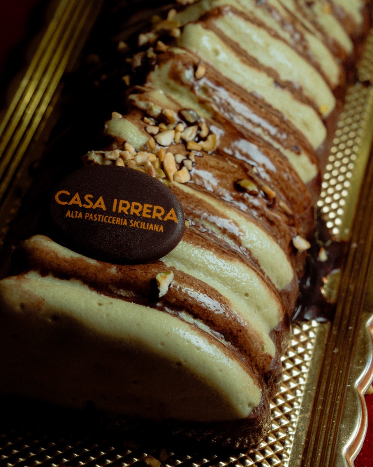 Sliced pistachio and chocolate log cake in layers, on a gold tray with a Casa Irrera branded disc