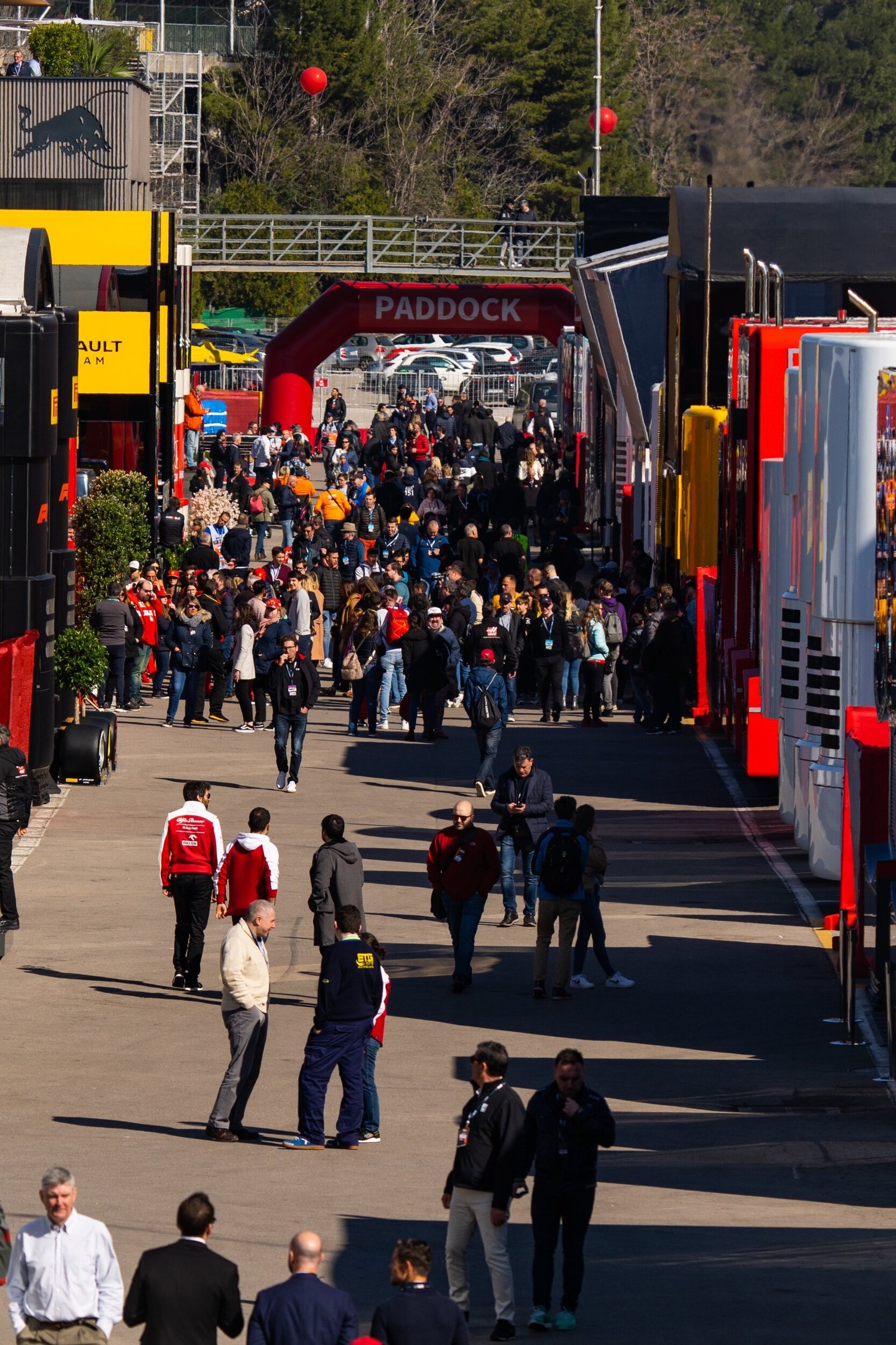 Formula 1 paddock at the Circuit de Barcelona-Catalunya during 2020 pre-season testing, crowds between the team hospitality units beneath the PADDOCK arch