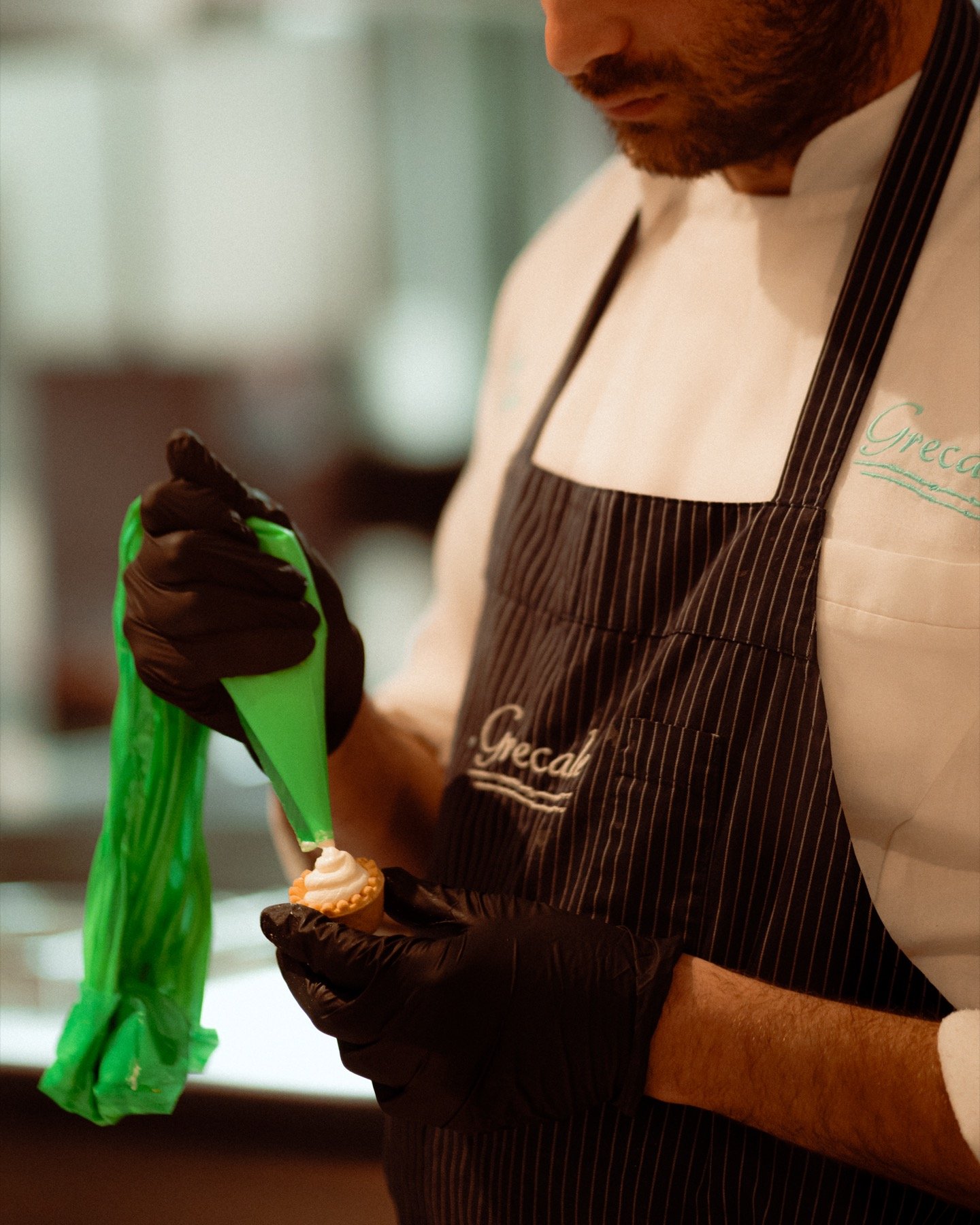 Grecale pastry chef piping cream onto a small tart, wearing branded apron