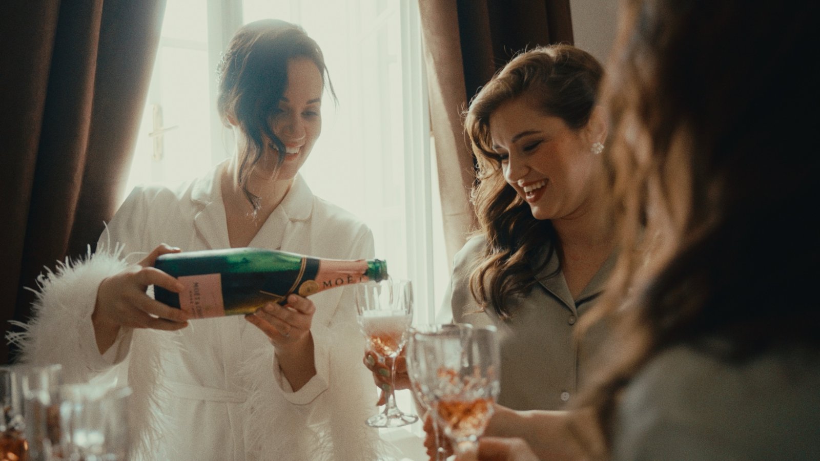 La sposa in vestaglia con bordo di piume versa champagne con una damigella nella luce della finestra