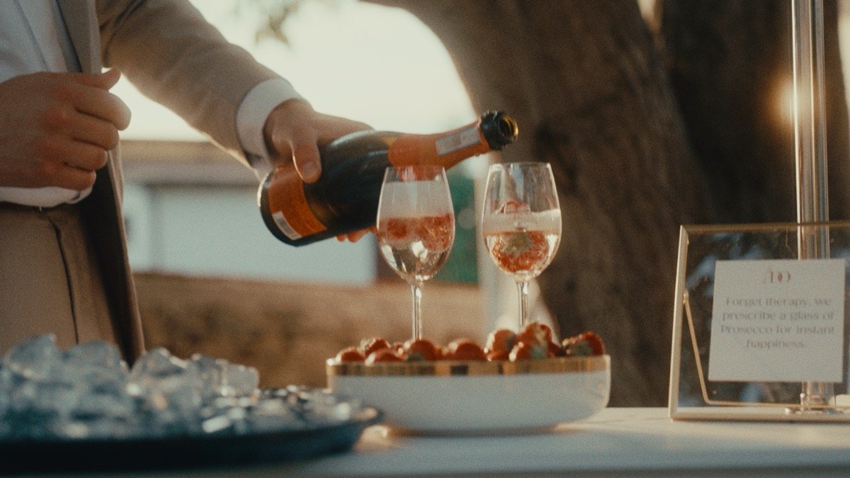 A guest pouring prosecco into two glasses over a bowl of strawberries, golden hour light