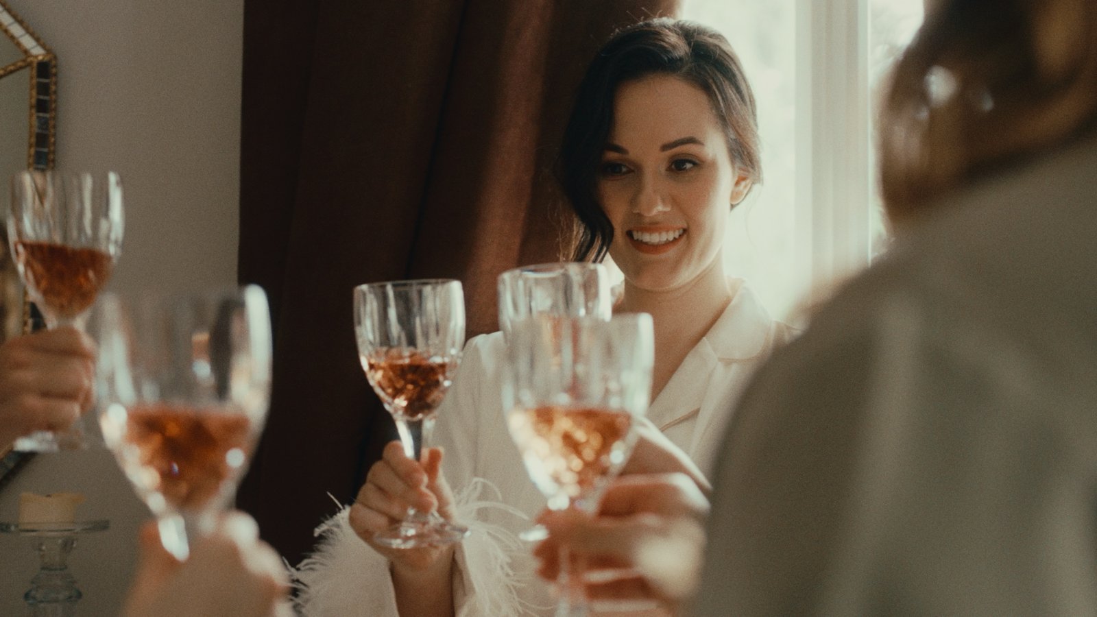 Calici di ros&eacute; alzati per un brindisi, la sposa sorride dietro, nella luce della finestra