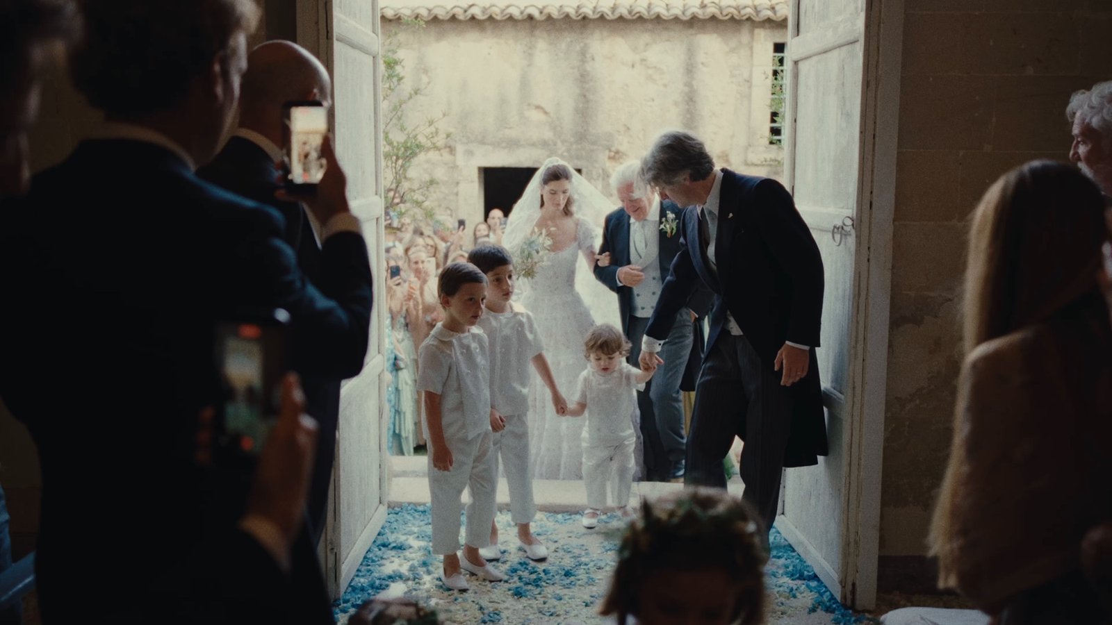 The bride entering on her father's arm, flower girls ahead of her and guests filming