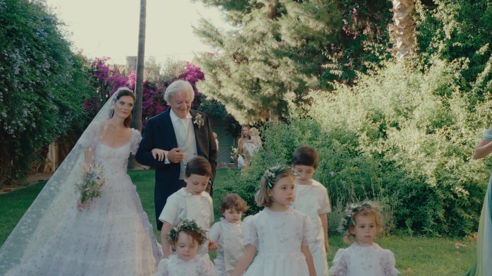 The bride walking through the garden with her father and the flower girls before the ceremony