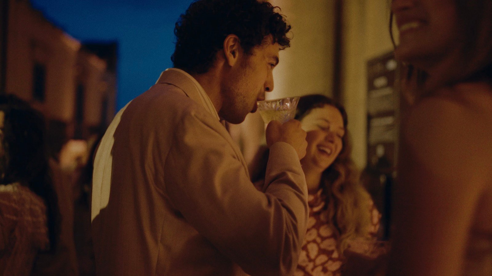 A guest drinking from a glass beside a laughing woman on a dusk street