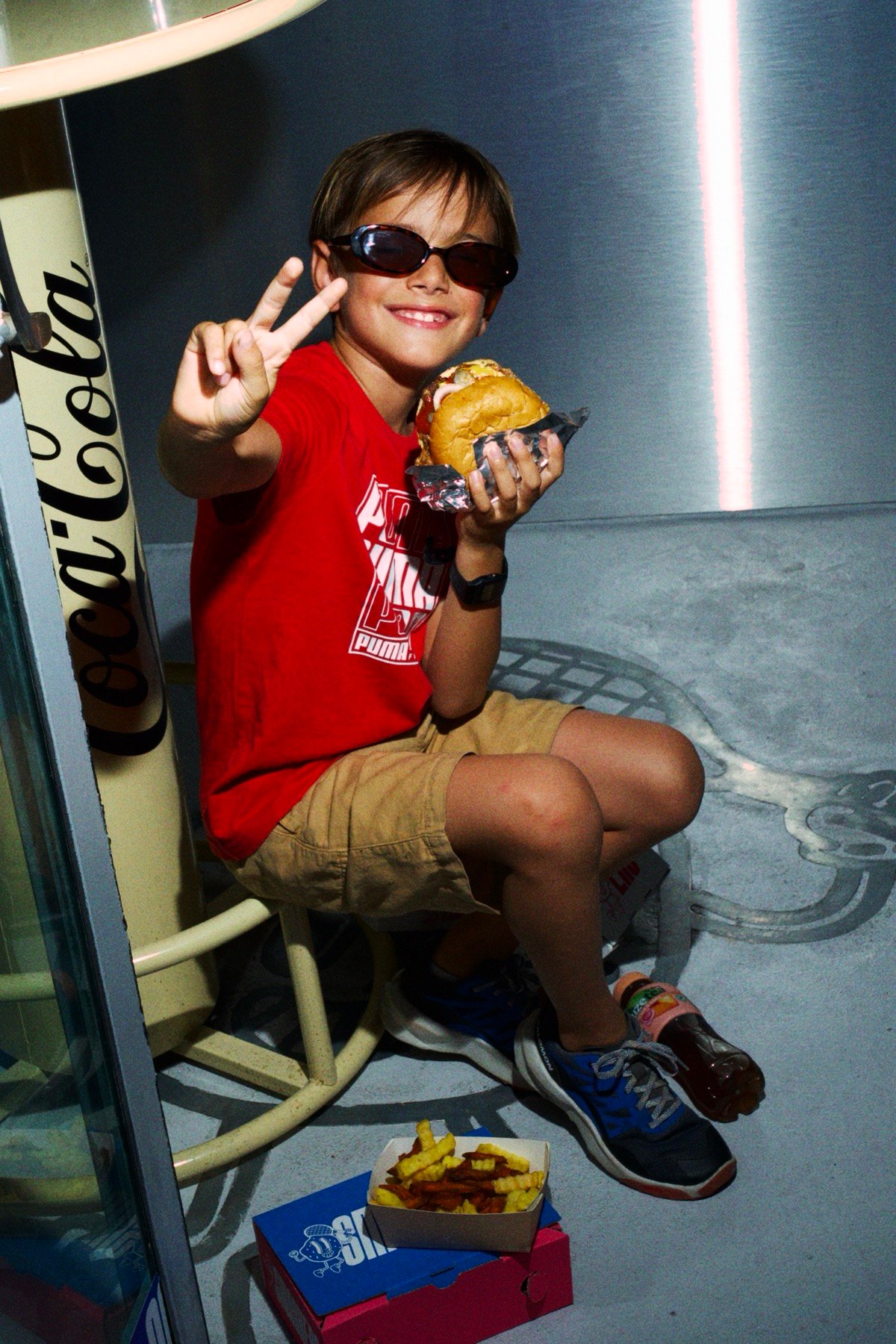 A boy in sunglasses holding up a peace sign with a Smashers burger in his other hand, fries and a drink on the floor beside him
