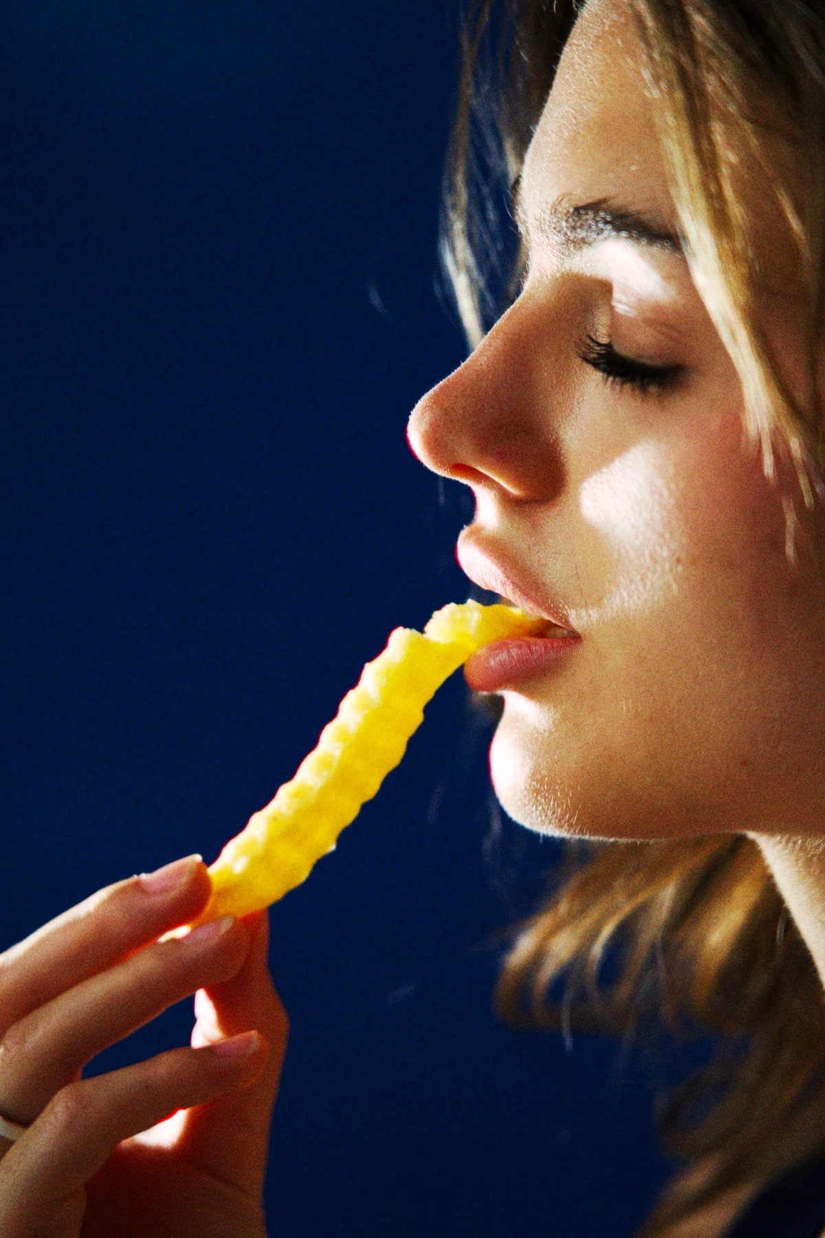 Close-up profile of a woman biting into a crinkle-cut fry, lit against a dark blue background