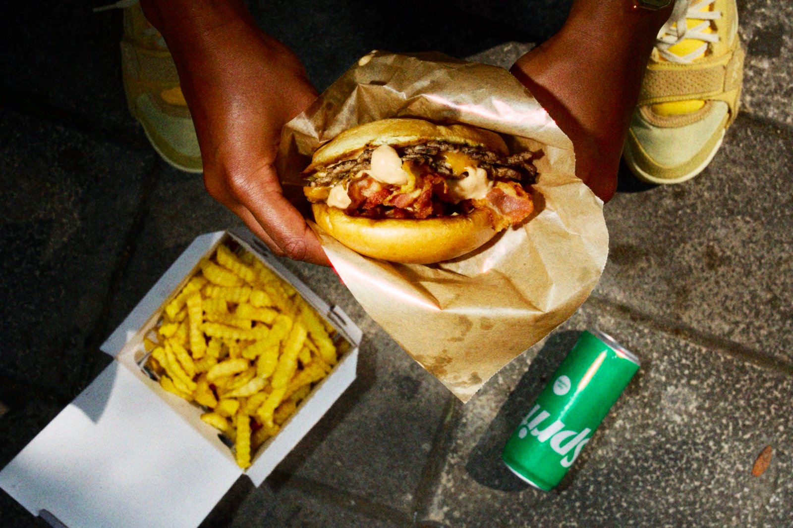 Overhead shot of hands holding a smashed double cheeseburger over a box of crinkle-cut fries, with a can of Sprite beside it