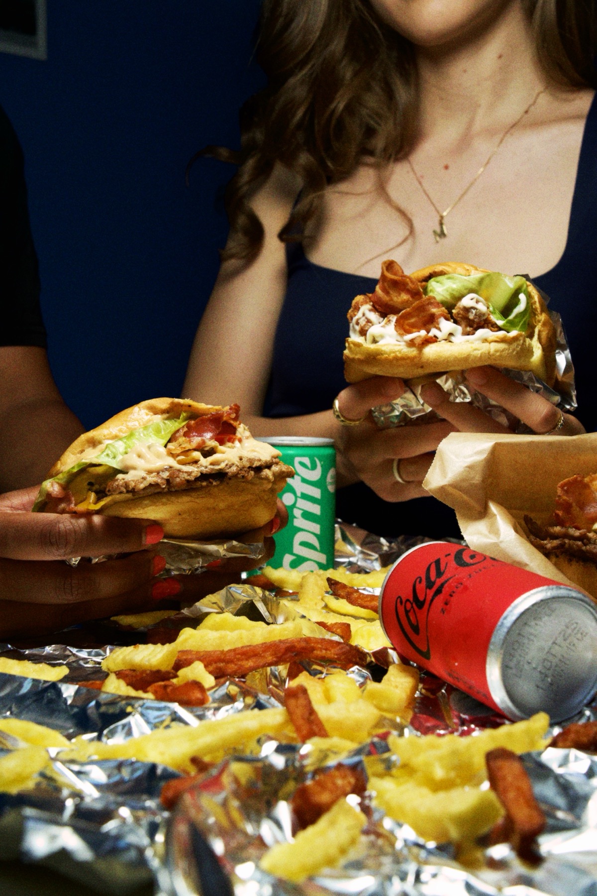 Two people holding smashed burgers over a table covered in fries, a can of Sprite and a can of Coca-Cola Zero