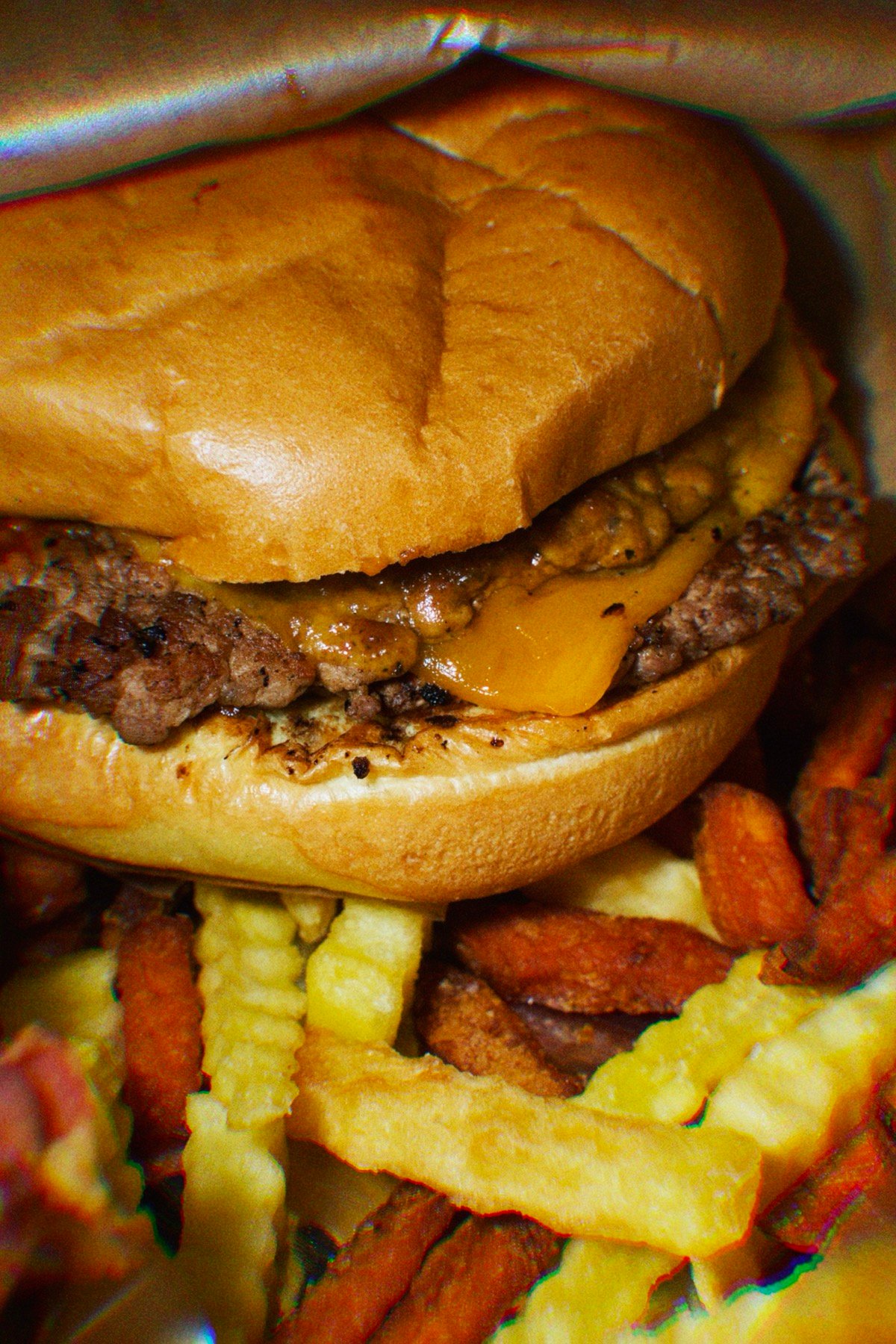 Extreme close-up of a Smashers double cheeseburger with melted cheese, stacked over a bed of crinkle-cut fries and bacon