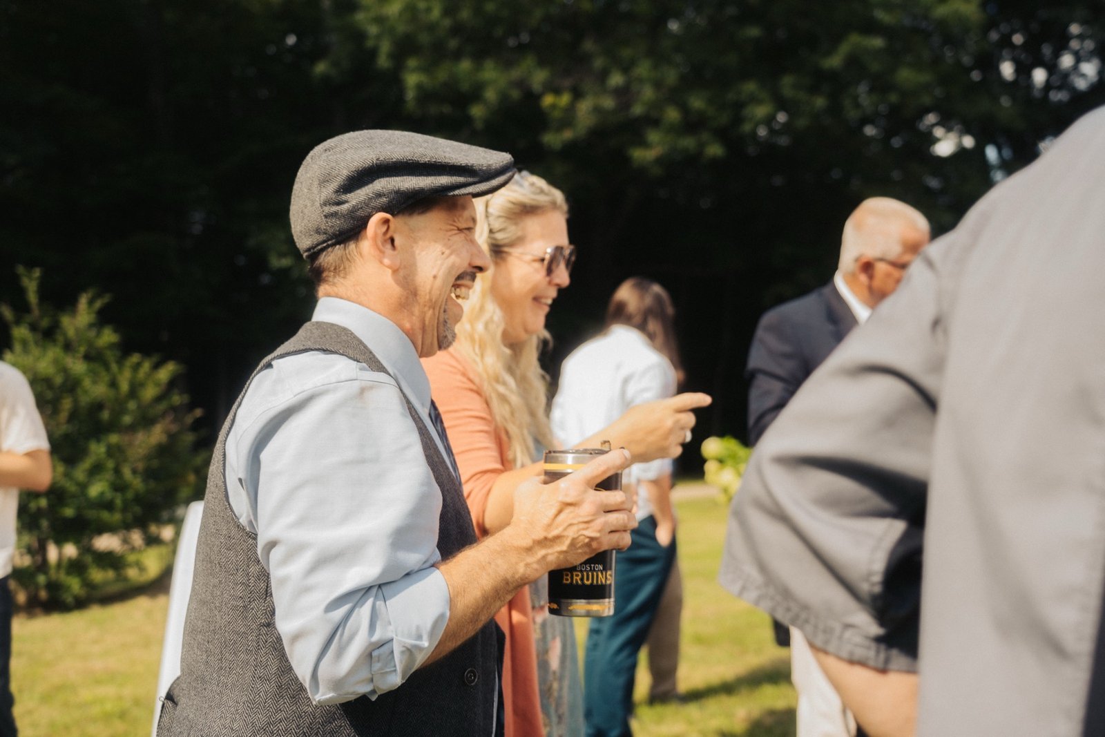 A wedding guest in a flat cap laughing candidly next to another guest in the evening light