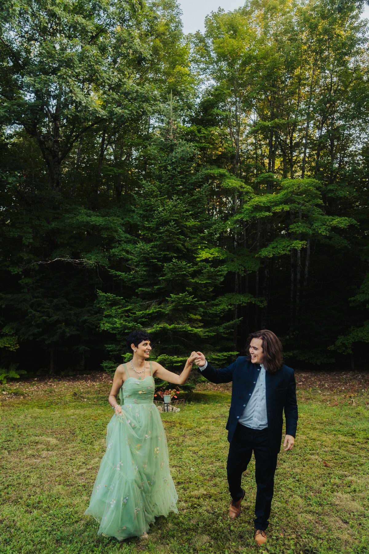 Bride in a pale green dress and groom holding hands, walking through a forest clearing