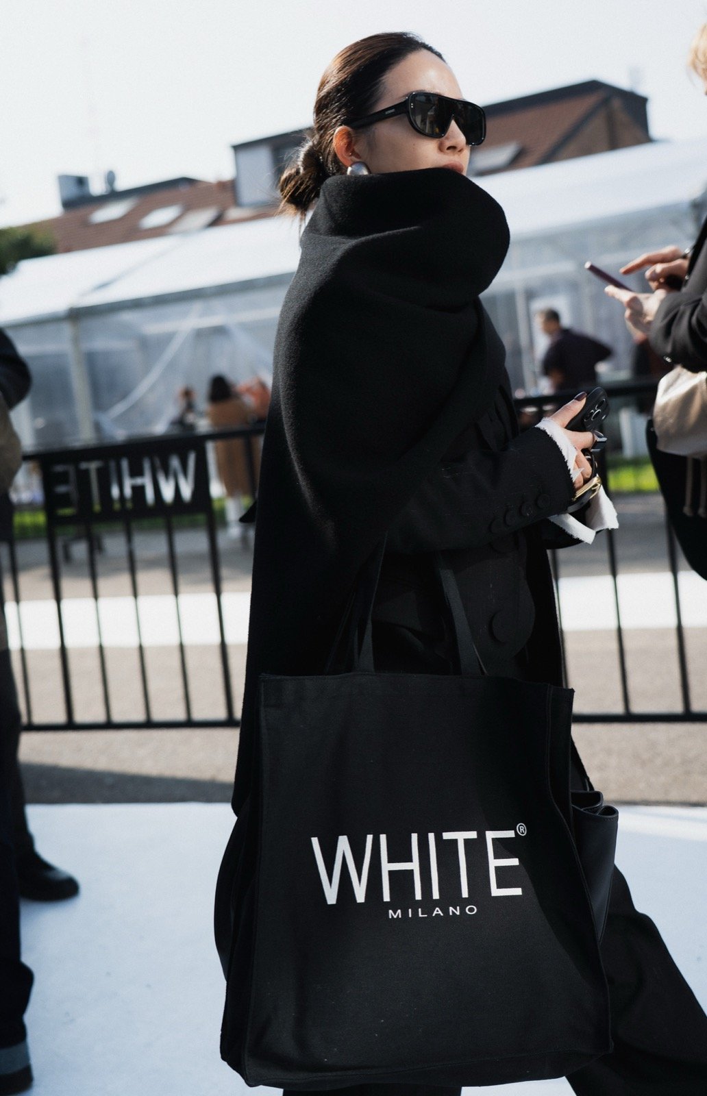 Attendee in an oversized black coat carrying a White Milano tote bag outside the show tent